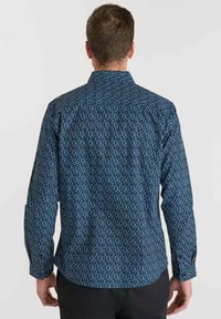 Man in een donkerblauw overhemd met lange mouwen en een klein wit abstract patroon, gezien vanaf de achterkant tegen een effen achtergrond.