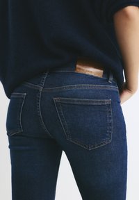Temno modri kavbojke iz denima z gladko teksturo, ki imajo rjav usnjen logotip na pasu ter žepoma na hrbtu z kontrastnim šivanjem.