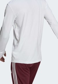 Man i långärmad vit strukturerad sportskjorta och vinröda träningsbyxor med vita ränder, sedd bakifrån.