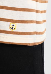 Pruhovaná košile s krátkým rukávem v krémové a hnědé barvě. Na lemu má malé vyšívané žluté kotvu. Měkká tkanina s uvolněným střihem.