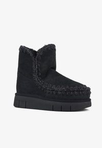Stivale alla caviglia in suede nero con cuciture texturizzate attorno alla parte superiore, dotato di una suola in gomma spessa e di una fodera morbida. Design semplice con accenti minimi.