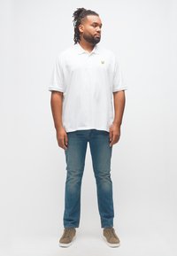 Άνδρας με dreadlocks που φοράει λευκό πόλο πουκάμισο, μπλε τζιν και μπεζ αθλητικά παπούτσια, στέκεται και κοιτάζει προς τα πλάγια σε απλό λευκό φόντο.