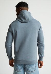 Sudadera gris con capucha y cordón, de mangas largas. Hecha de material suave, presenta puños acanalados y un diseño liso. Combinada con jeans de mezclilla clara.