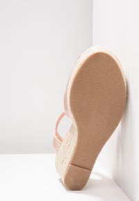 Vue de la semelle d'une sandale compensée rose à paillettes avec semelle en caoutchouc texturée et plateforme en jute tissée, positionnée contre des murs d'angle blancs.