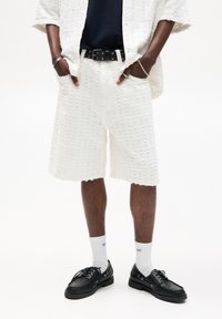 Shorts bianchi con una texture e una vestibilità ampia, abbinati a una maglietta blu navy, una cintura nera e scarpe nere. I dettagli notevoli includono un motivo a pois e orli cuffed.