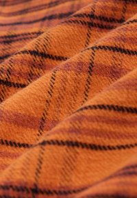 Oranje stof met een zachte textuur, voorzien van een ruitpatroon in zwart en paars tinten, gerangschikt in diagonale lijnen over het oppervlak.