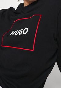 Sudadera negra con un contorno rectangular rojo, que presenta el nombre de la marca en blanco "HUGO" de manera destacada en el centro. Textura suave.