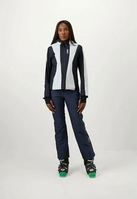 Σακάκι σκι σε ναυτικό μπλε και ανοιχτό γκρι με φερμουάρ μπροστά, εφαρμοστή σχεδίαση και αντίθετα πάνελ. Συνδυασμένο με ναυτικές παντελόνες σκι και μαύρες μπότες.