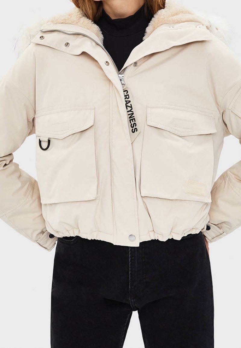 Veste courte beige avec grandes poches avant, col en fausse fourrure, tirette de fermeture éclair étiquetée "CRAZINESS,", portée sur un pantalon et un top noirs.