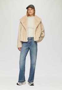 Faux-Fur-Crememantel mit breitem Kragen, geripptem hellen Oberteil, blauen Bootcut-Jeans und grauem Cap; silberne Sneakers mit strukturiertem Finish.