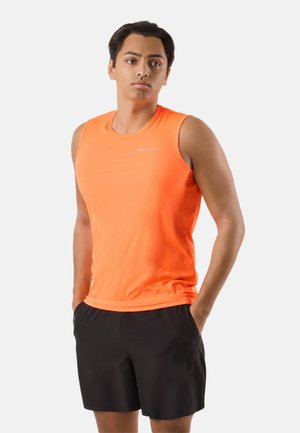 Giovane uomo con maglietta sportiva arancione senza maniche e pantaloncini neri, in piedi con le mani in tasca, sfondo neutro.