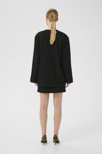 Černá tvídová bunda s dlouhými rukávy, texturovaným vzorem a jemným leskem, v kombinaci s odpovídající mini sukní. Obuta v tmavě zelené barvě.