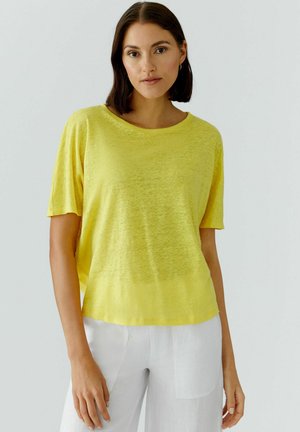 Frau mit schulterlangem dunklem Haar, die ein locker sitzendes gelbes Kurzarmtop und weiße Hosen trägt, vor einem einfarbigen Hintergrund stehend.