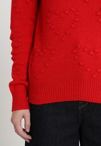 Pull en maille rouge présentant un motif texturé avec des points en relief, des poignets et un ourlet côtelés. Associé à un jean en denim foncé.