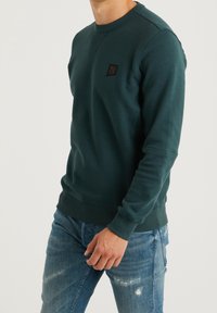 Tmavě zelená mikina s kulatým výstřihem, ribovanými manžetami a malou černou nášivkou na hrudi; nošena s opotřebovanými modrými džíny.