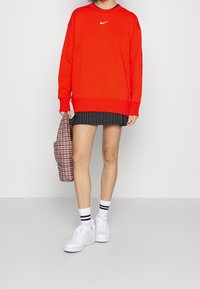 Roter Sweatshirt mit einem weißen Nike-Logo, kombiniert mit einem schwarz gestreiften Rock, weißen Sneakers und einer karierten Tasche. Lässiger sportlicher Stil.