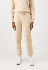 Lichtbeige sweatpants met elastische boorden, trekkoord in de taille en een klein zwart logo detail aan de linkerkant. Zachte stofstructuur.