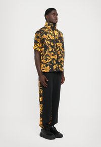 Homme portant un pantalon noir et des baskets noires avec une veste à manches courtes noire et jaune à motifs, se tenant contre un fond blanc.