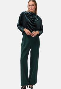 Samtiger, drapierter grüner Oberteil mit langen Ärmeln, kombiniert mit hochgeschnittenen, weit geschnittenen grünen Hosen. Akzentuiert mit verzierten schwarzen Absatzschuhen.