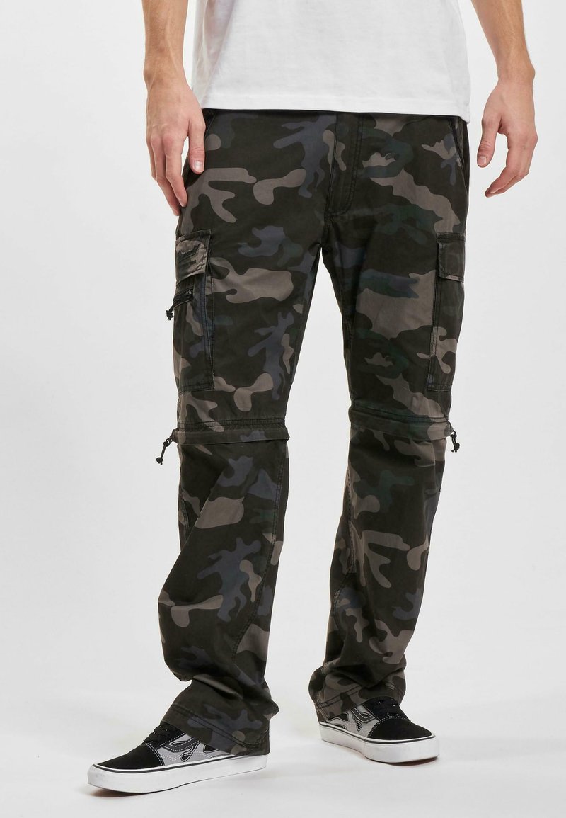 Homme portant un pantalon cargo camouflage noir et gris avec plusieurs poches et des baskets noir et blanc, se tenant devant un fond uniforme.