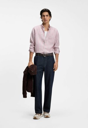 Ung mand iført rød- og hvidstribet skjorte over en hvid t-shirt, mørke jeans med sort bælte, beige sneakers, holder en mørk jakke i venstre hånd.