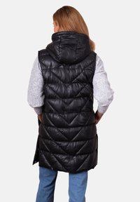 Woman with light brown hair wears black quilted sleeveless hooded vest over gray sweater and blue jeans, facing away from camera.