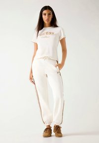 T-shirt en coton blanc avec le logo "GUESS", associé à un pantalon de jogging crème avec des bandes latérales marron, des poignets élastiques et une taille à cordon. Baskets marron.