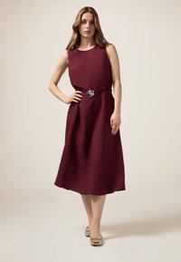 Rochie midi burgundă cu o textură netedă și design fără mâneci, având o talie gathered și un accent decorativ sub formă de broșă argintie.