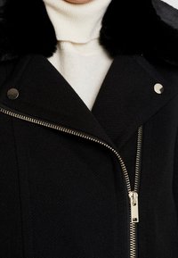 Chaqueta negra con una tela texturizada, presenta un cuello de piel sintética, herrajes plateados y un cierre de cremallera diagonal en el frente.