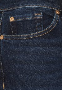 Jeans de mezclilla azul oscuro con un bolsillo delantero que presenta costuras doradas contrastantes y herrajes de metal. Textura suave con un ligero brillo.