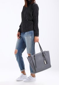 Sac fourre-tout en cuir gris avec deux poignées supérieures, design minimaliste, avec une étiquette suspendue. Porté avec un haut rayé noir et un jean usé.