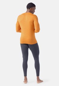 Maglietta arancione a maniche lunghe e leggings grigi, design aderente, texture morbida, tessuto liscio, dettagli di cucitura minimi, indossata a piedi nudi.