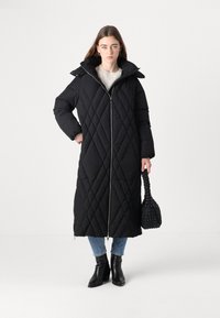 Crni duguljasti punjeni kaput s visokim ovratnikom, prednjim patentnim zatvaračem i bočnim prorezima, nosi se uz plave traperice i crne čizme s potpeticom. Drži crnu torbu.