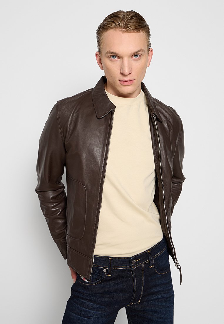 Veste en cuir marron avec un col classique, fermeture éclair devant, poches latérales et une coupe ajustée, associée à une chemise légère et un jean foncé.