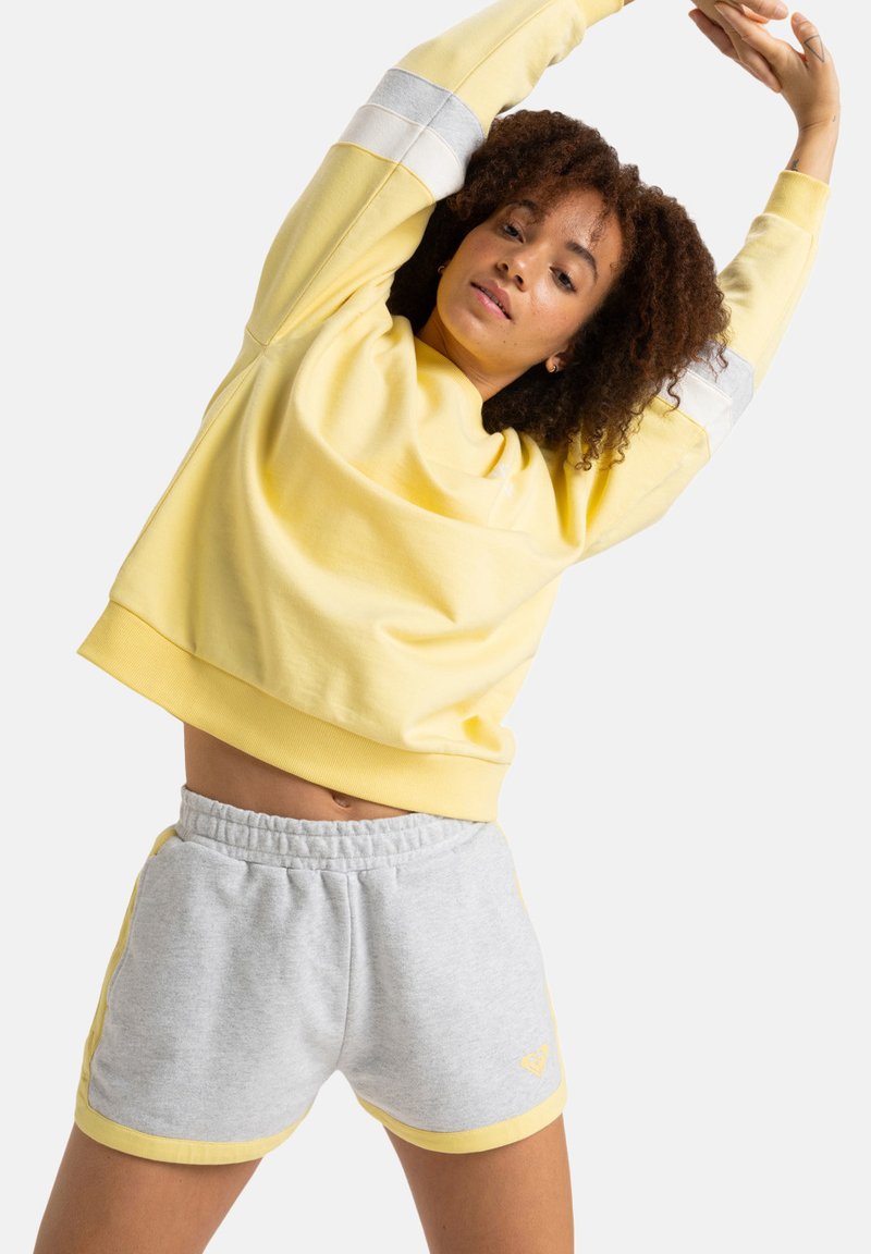 Sudadera amarilla con rayas grises en las mangas, combinada con pantalones cortos grises con acentos amarillos. Material de algodón suave, corte relajado.