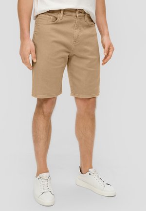 Homme portant un short beige jusqu'aux genoux, des baskets blanches et une chemise blanche sur un fond uni.