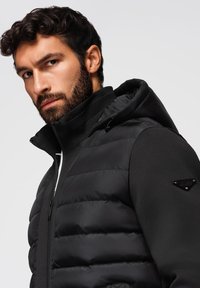 Uomo con capelli ricci scuri e barba, che indossa una giacca imbottita nera con cappuccio e colletto alto, mentre guarda leggermente di lato.