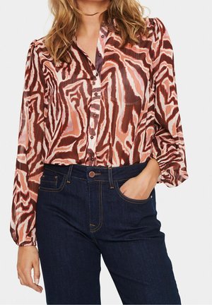 Vrouw met een blouse met lange mouwen en knopen, met een abstract patroon in rood, roze en bruin, gestopt in donkerblauwe spijkerbroek. Eén hand in de zak.