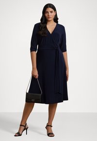 Abito avvolgente blu navy con maniche a tre quarti, dettaglio della cintura annodata e gonna fluida. Abbinato a una borsa a tracolla nera con texture e tacchi a strappo.