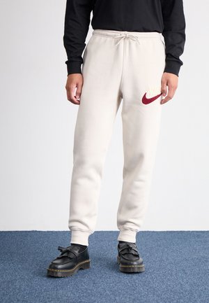 Person iført cremefarvede joggingbukser med et rødt Nike-logo, sort langærmet shirt og sorte lædersko med kvaster på blåt tæppe.