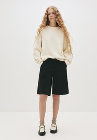 Jasnobeżowy, oversize sweter z prążkowanym dołem, w połączeniu z czarnymi szortami do kolan. Modelka ma na sobie taupe skarpetki oraz białe buty z czarnymi detalami.