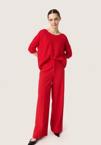 Rød strikket sweater med korte ærmer, parret med højtaljede vide røde bukser og sorte spidse sko, på en neutral baggrund.