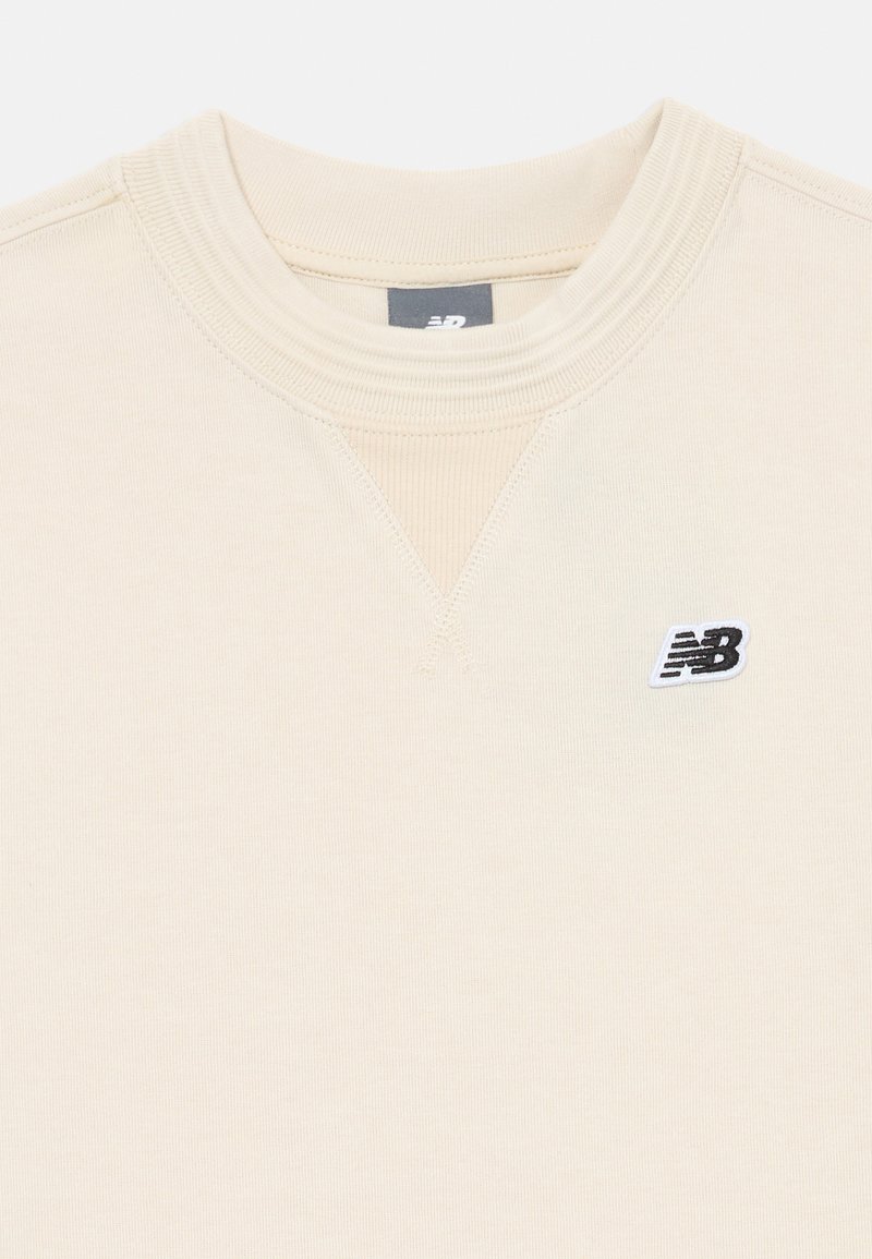 Sudadera beige con cuello acanalado y costuras texturizadas, y un pequeño logotipo bordado en blanco y negro en el lado izquierdo.