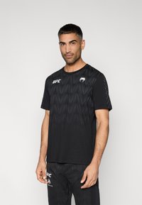 Μαύρο αθλητικό μπλουζάκι με γεωμετρικό σχέδιο, κοντά μανίκια και λογότυπα UFC στο εμπρός μέρος. Μαλακό ύφασμα με απαλή υφή.