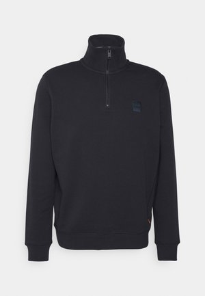 Sudadera negra con cuello alto y cierre de media cremallera. Presenta puños de canalé y un pequeño parche con el logo en el pecho. Tejido suave y texturizado.