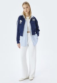 Chaqueta azul con cremallera y rayas blancas, superpuesta sobre una camisa a rayas azul y blanca, combinada con pantalones blancos y zapatos de color claro.