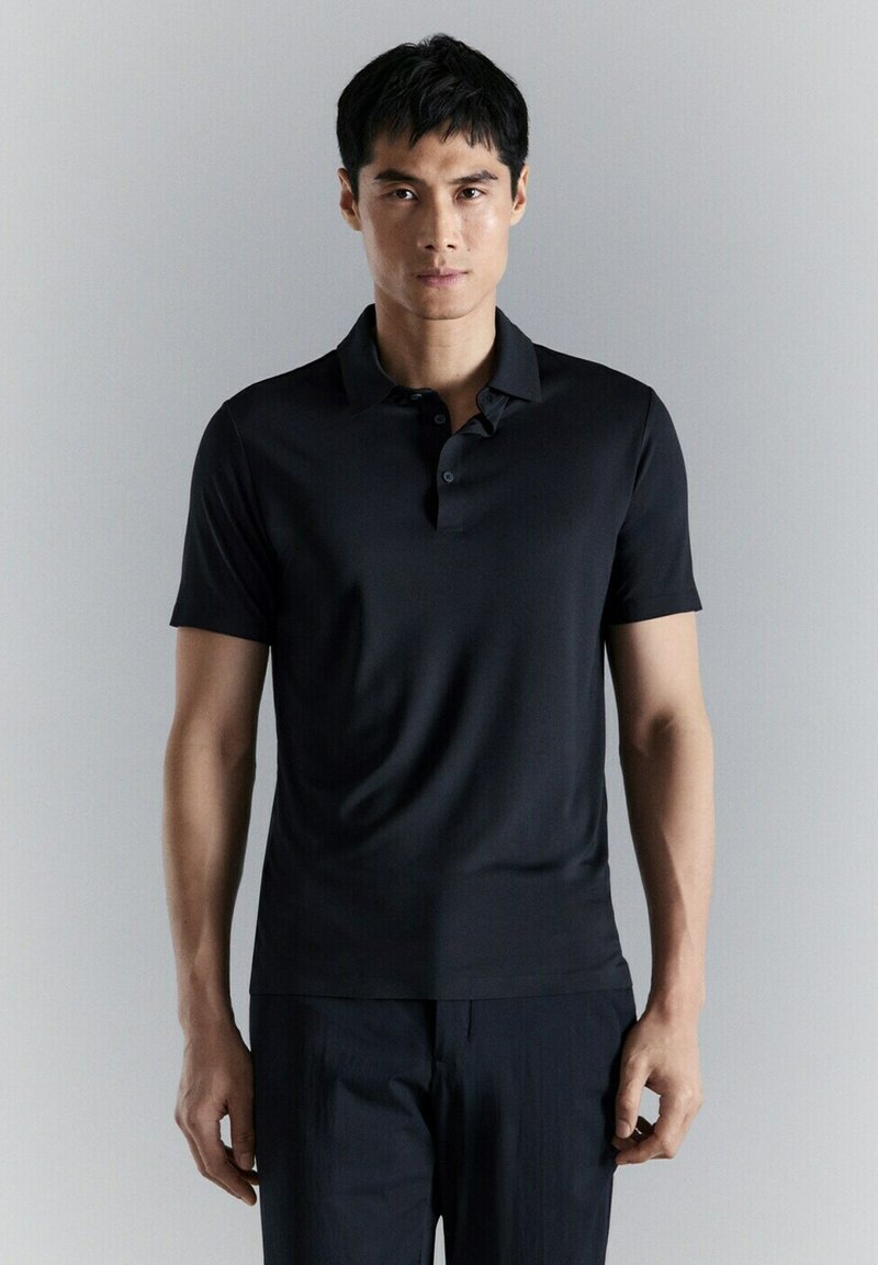 Jeune homme debout devant un fond gris clair uni, portant un polo noir ajusté à manches courtes et un pantalon noir.