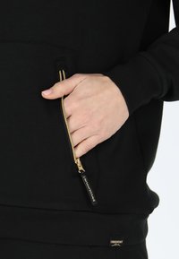 Svart sweatshirt med en ficka fram med en guldfärgad dragkedja. Texturen ser slät ut, och den har en liten logotyp nertill.