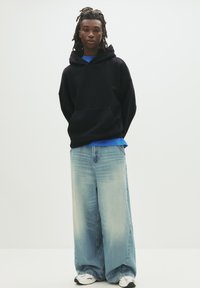 Sort oversized hættetrøje, blå lagdelt t-shirt under, lyseblå brede jeans og hvide sneakers. Glat stoftekstur.