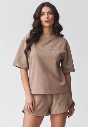 Kobieta z długimi ciemnymi włosami, ubrana w luźny beżowy t-shirt „MAKADAMIA” i pasujące krótkie spodenki, stoi na tle jednolitego jasnego tła.